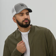 Man wearing a gray trucker cap with a mesh back and daily logo, dressed in a casual attire, outdoors.